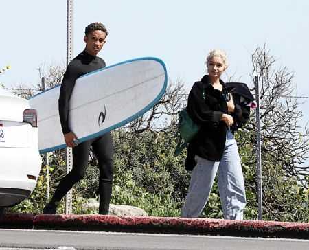 Jaden sosteniendo una tabla de surf y una chica rubia caminando junto a él.