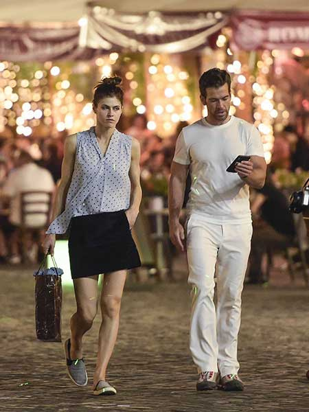   Alexandra Daddario (à esquerda) em uma camisa branca de bolinhas e shorts pretos e seu ex-namorado Brendan Wallace (à direita) em uma camiseta branca e calça creme