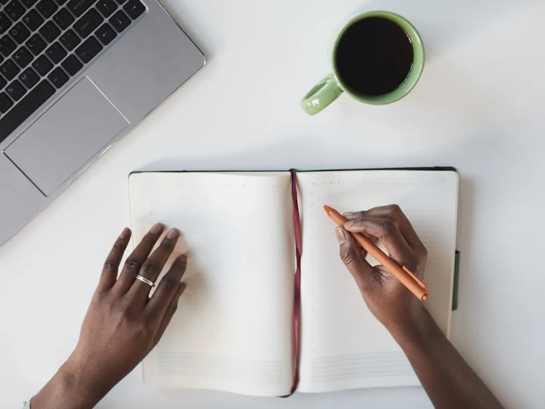 Minimal top view of unrecognizable African-American woman writing in planner at white business workplace' decoding='async' fetchpriority='high